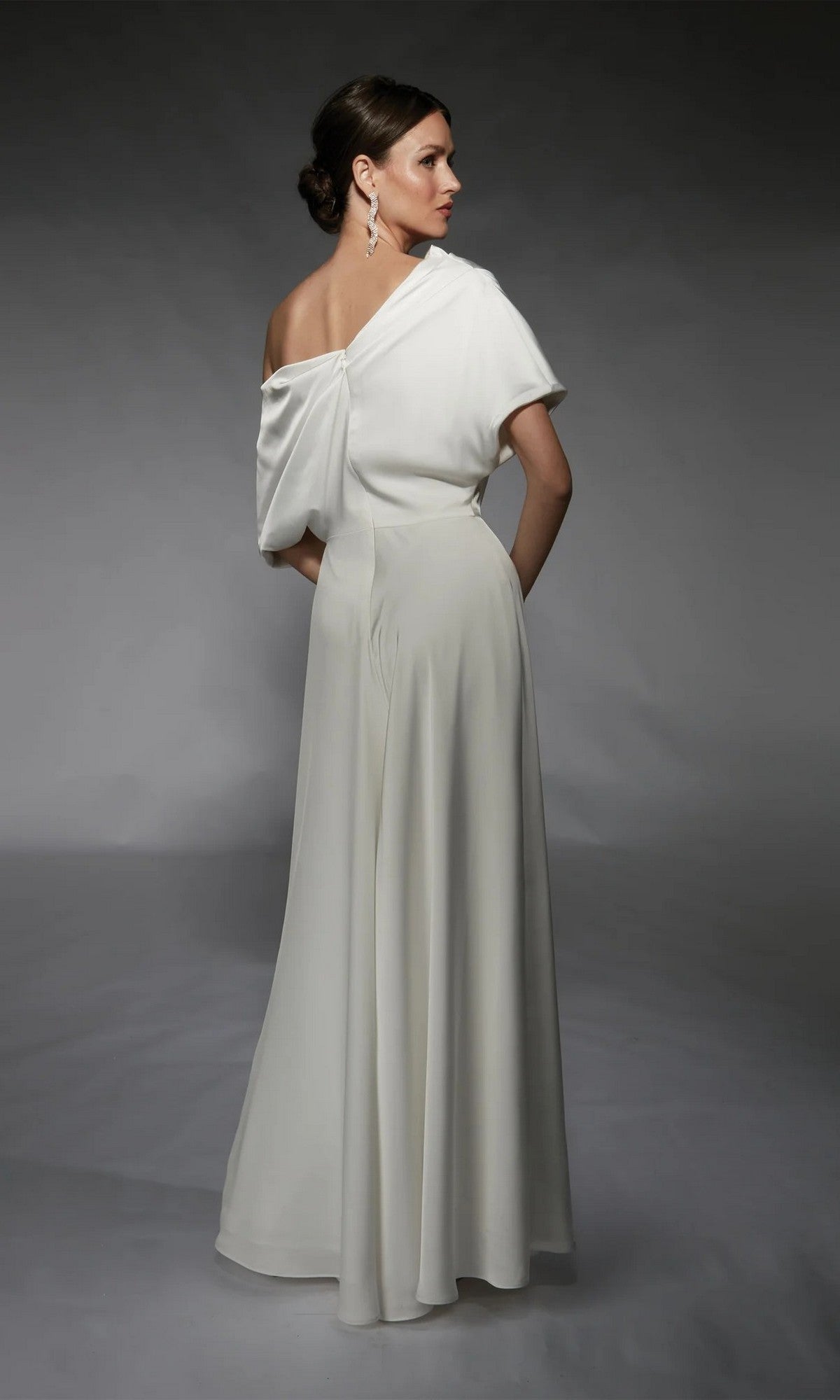 A woman faces away from the camera in the Alyce 70072, a long white satin jumpsuit with a one-shoulder, floor-length draped design, set against a neutral gray background.